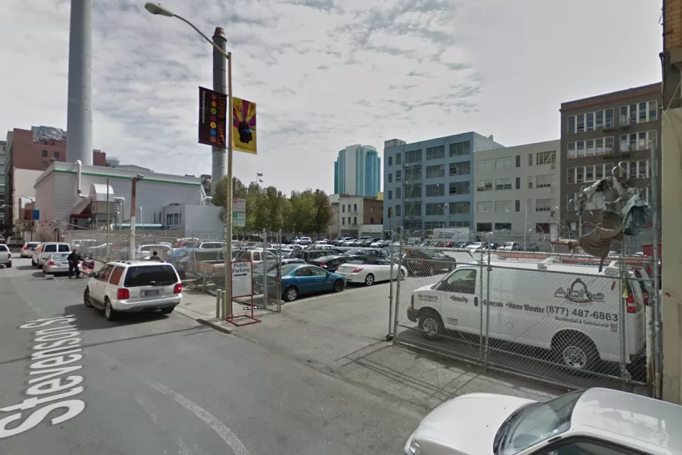 A surface parking lot, full of cars, is located between tall buildings and urban density.